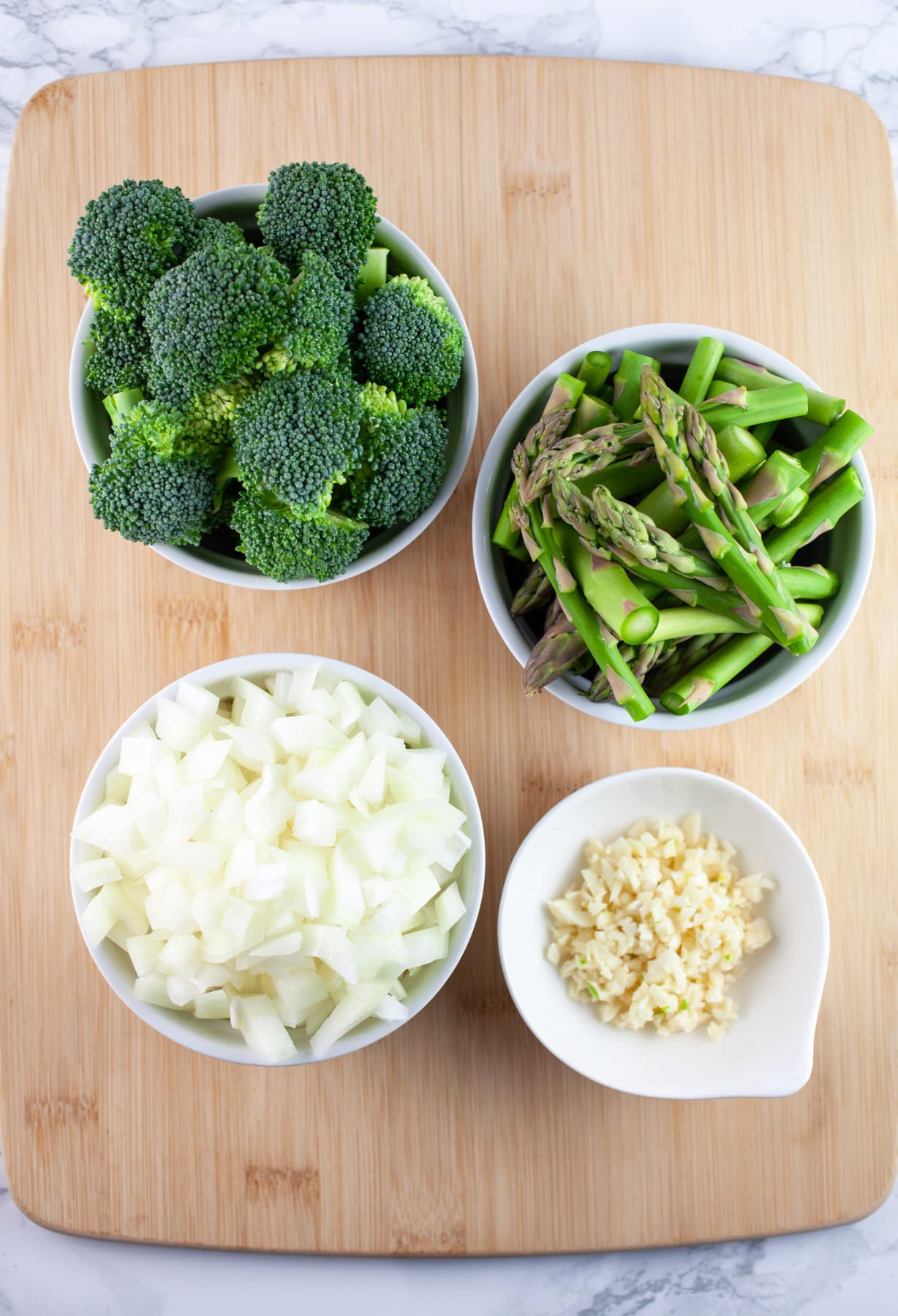 Minced garlic and onions, chopped asparagus and broccoli florets in small white bowls on wooden cutting board.