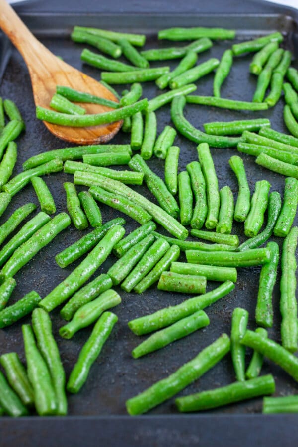 Raw green beans on baking sheet with wooden spoon.
