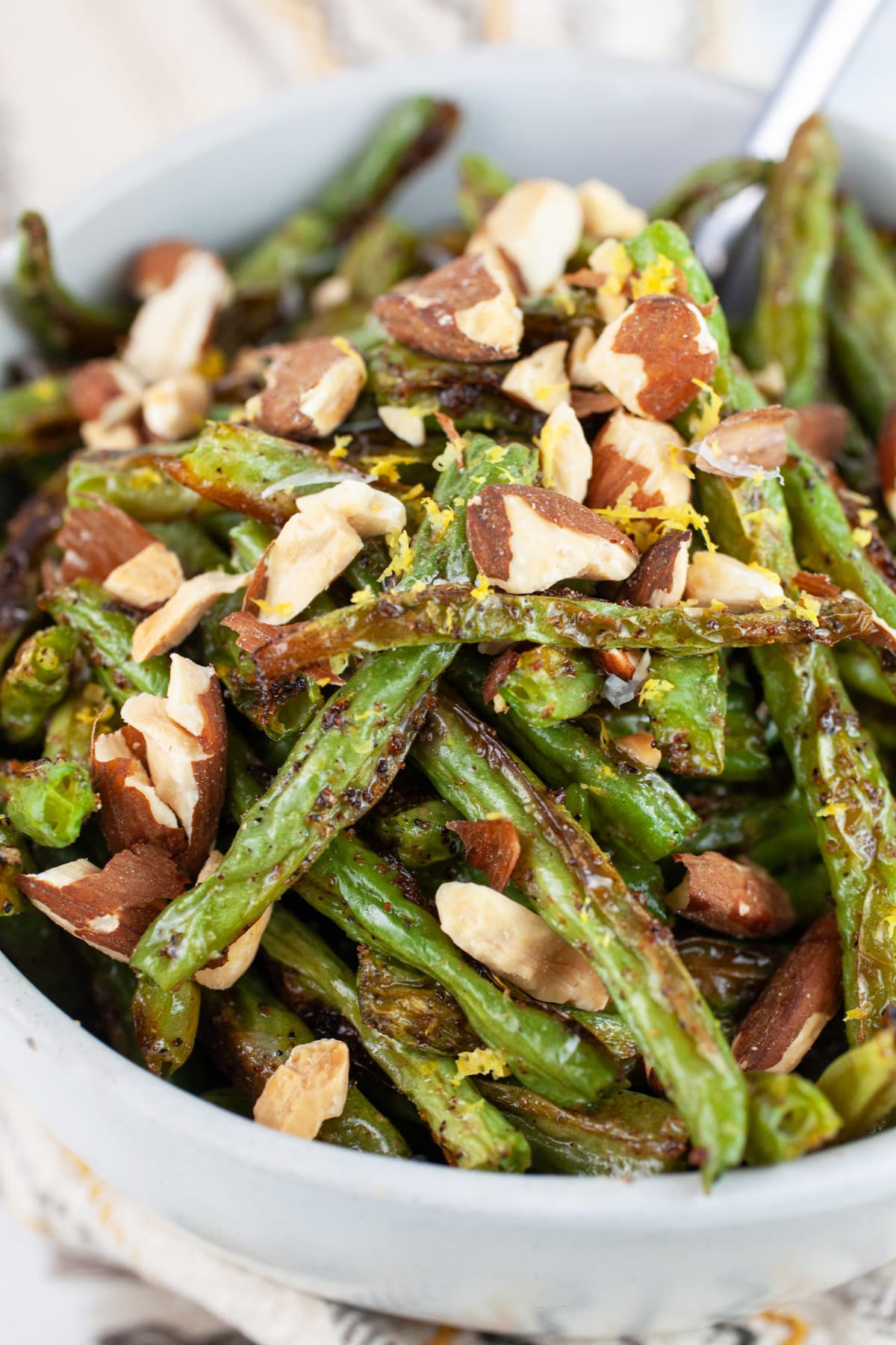 These Simple Garlicky Roasted Green Beans is one of the best easy and healthy side dishes out there! Fresh green beans are tossed in olive oil and garlic powder and then baked in the oven until nice and crispy and then topped with almonds. This quick vegetarian and vegan recipe is perfect for lunch or dinner! #greenbeans #vegetarian #vegan #garlic #withalmonds #easy Garlic roasted green beans with toasted almonds in white bowl with spoon.