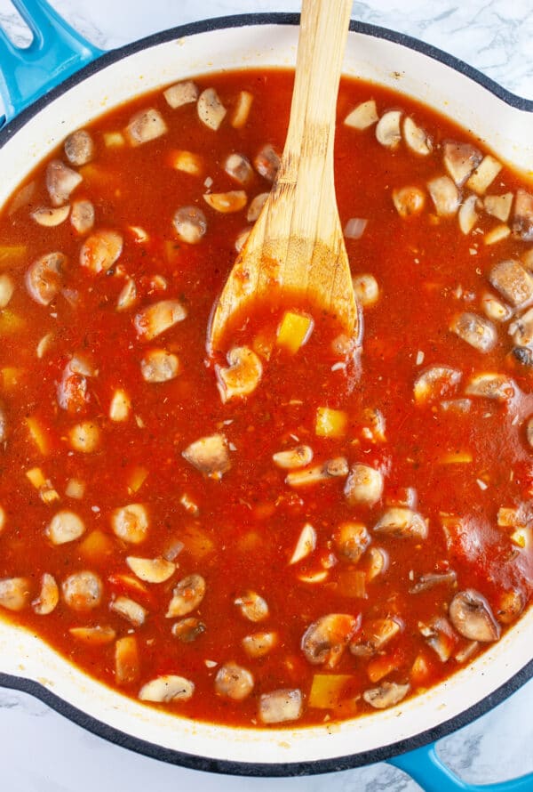 Mushrooms and bell peppers with marinara sauce in skillet with wooden spoon.