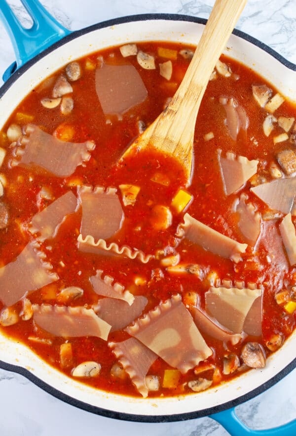 Uncooked lasagna noodles with marinara sauce in skillet with wooden spoon.