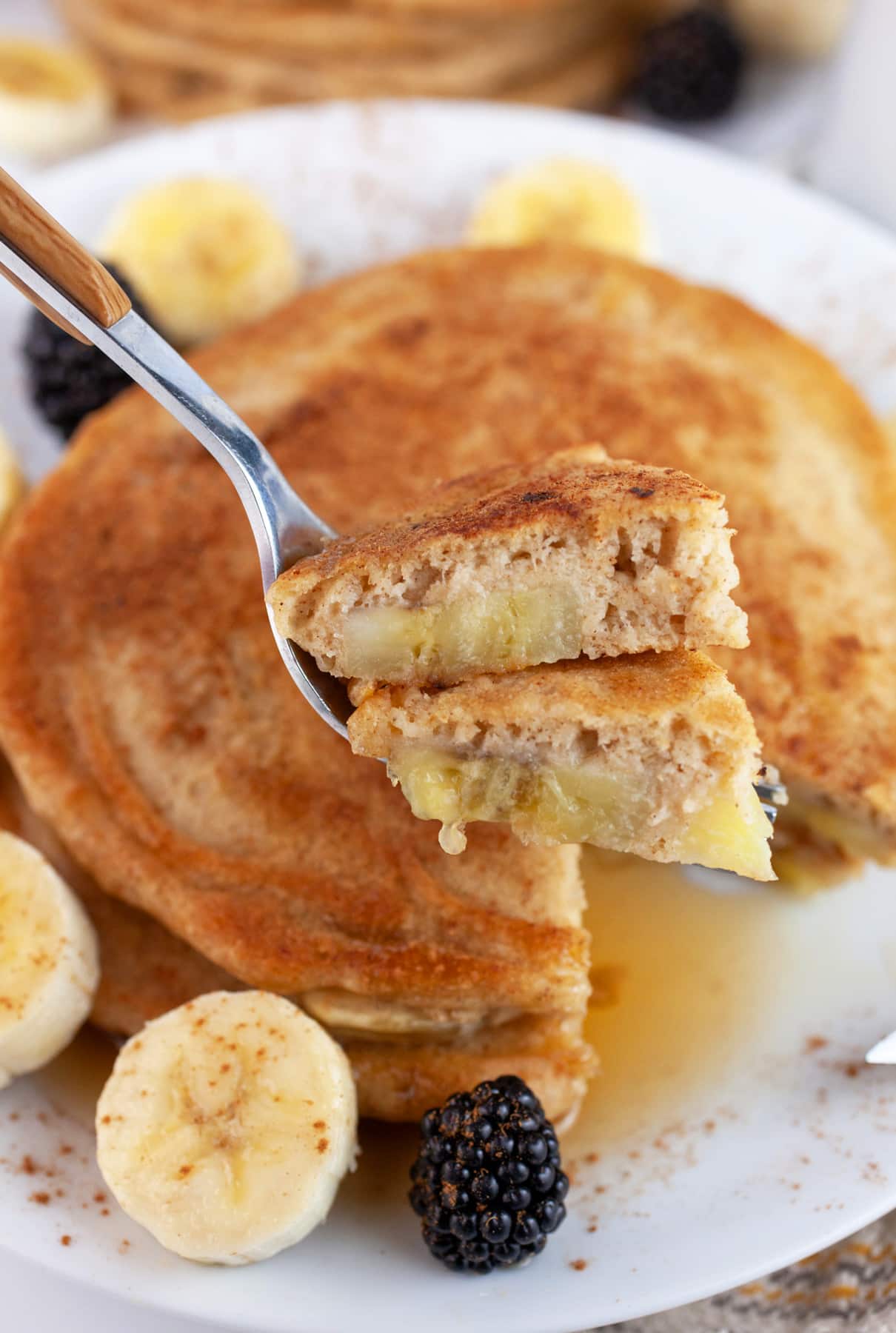 These Old-Fashioned Banana Pancakes are the perfect breakfast for weekday mornings or a weekend treat! They're light and fluffy, easy to make, and fun to eat. This simple pancake recipe is adapted from an old church cookbook. These banana pancakes are made from scratch and topped with maple syrup. Homemade is always best! #ad @kempscows #pancakes #banana #oldfashioned Forkful of banana cinnamon pancakes lifted from white plate.
