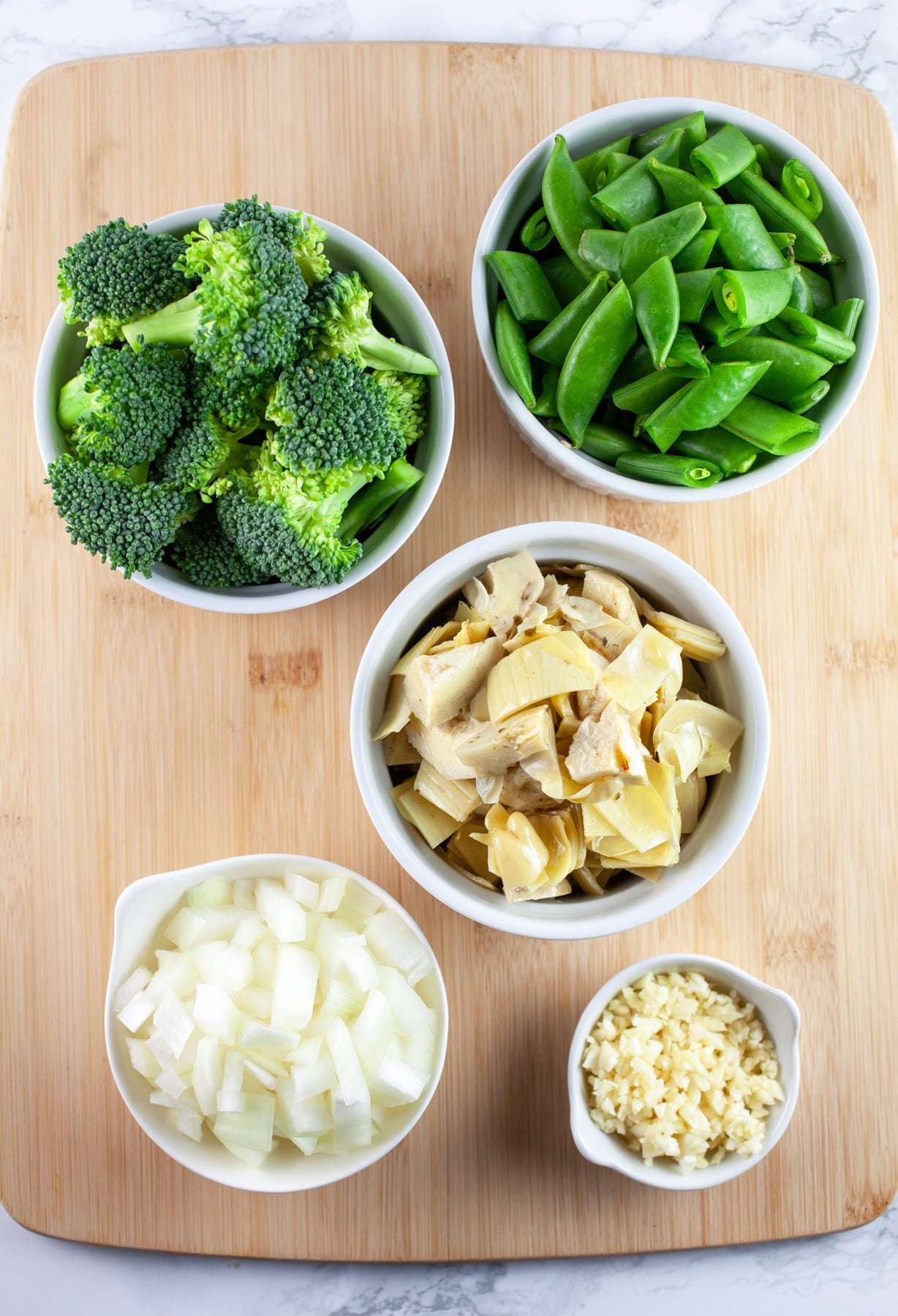 Minced garlic, onions, chopped artichoke hearts and snap peas, and broccoli florets in small white bowls on wooden cutting board.