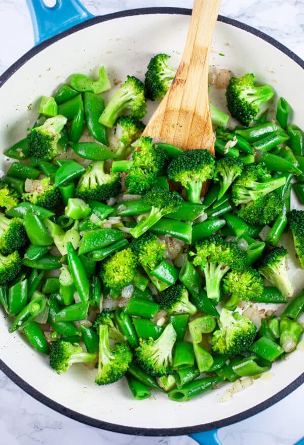 Snap peas and broccoli florets sauteed in skillet with wooden spoon.