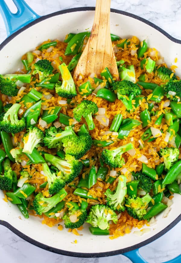 Snap peas and broccoli florets sauteed with uncooked orzo pasta in skillet with wooden spoon.