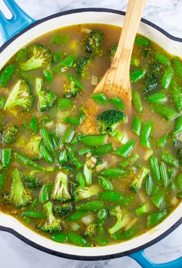 Snap peas and broccoli florets simmered in broth in skillet with wooden spoon.