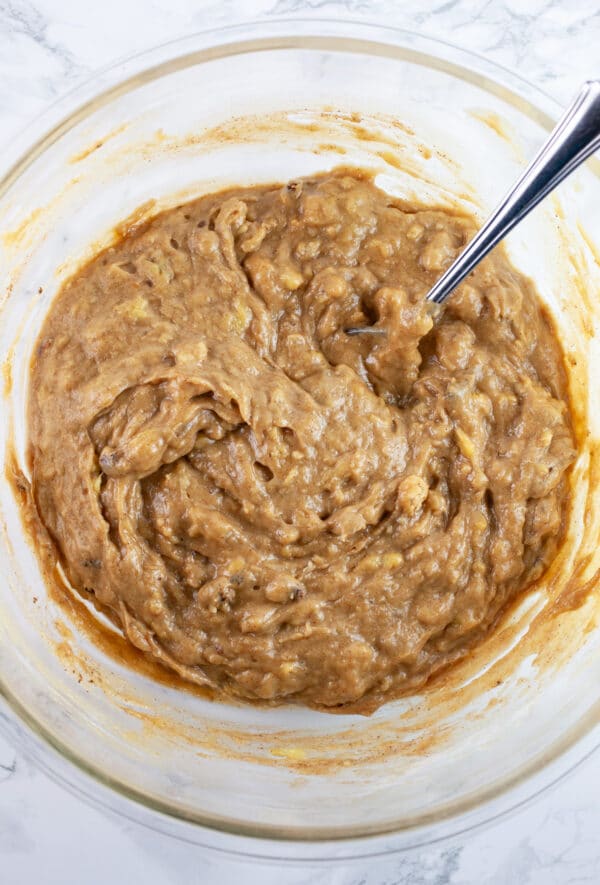 Banana muffin batter with cinnamon and toasted walnuts combined in glass mixing bowl with fork.