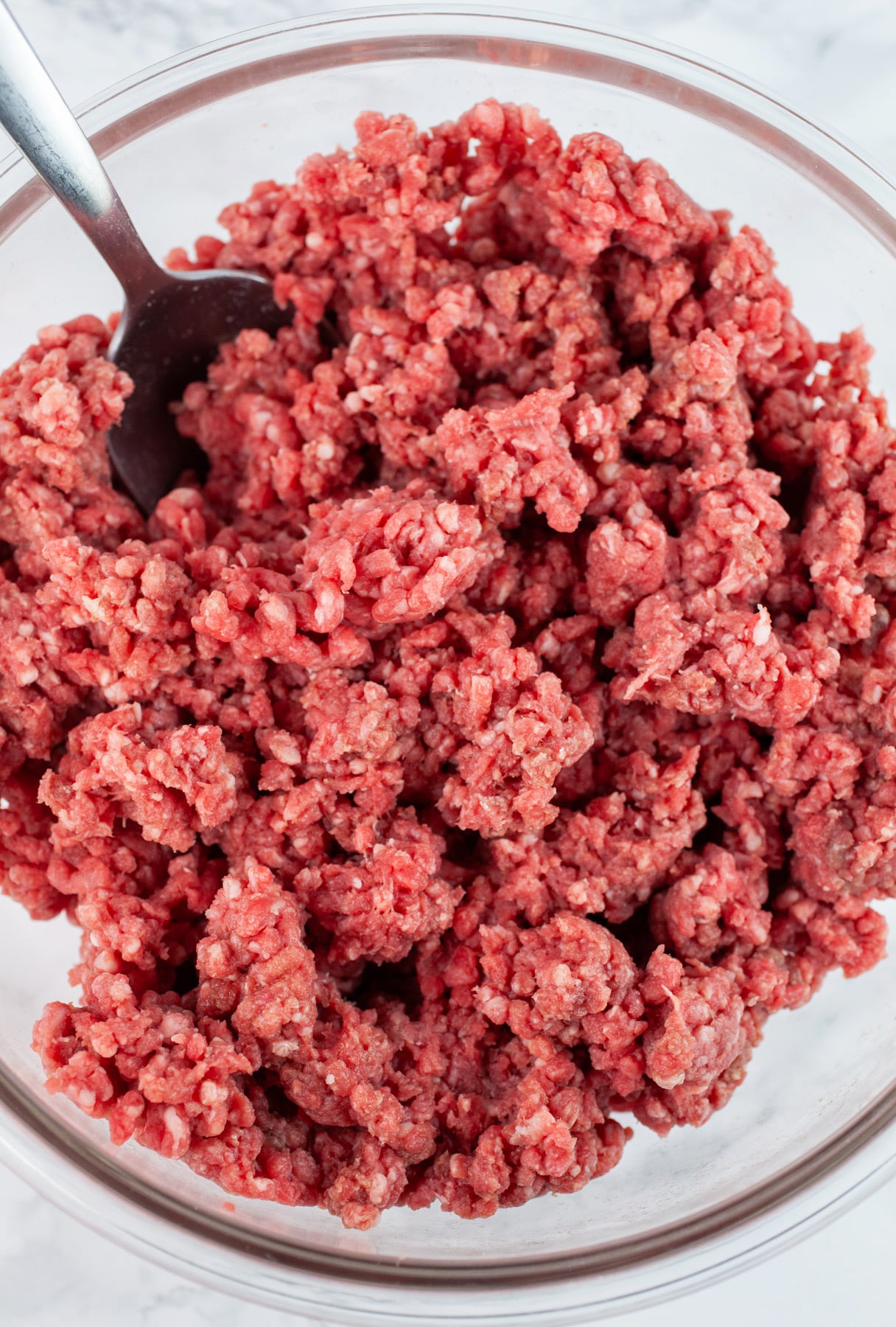 Raw ground beef in glass mixing bowl with metal spoon.