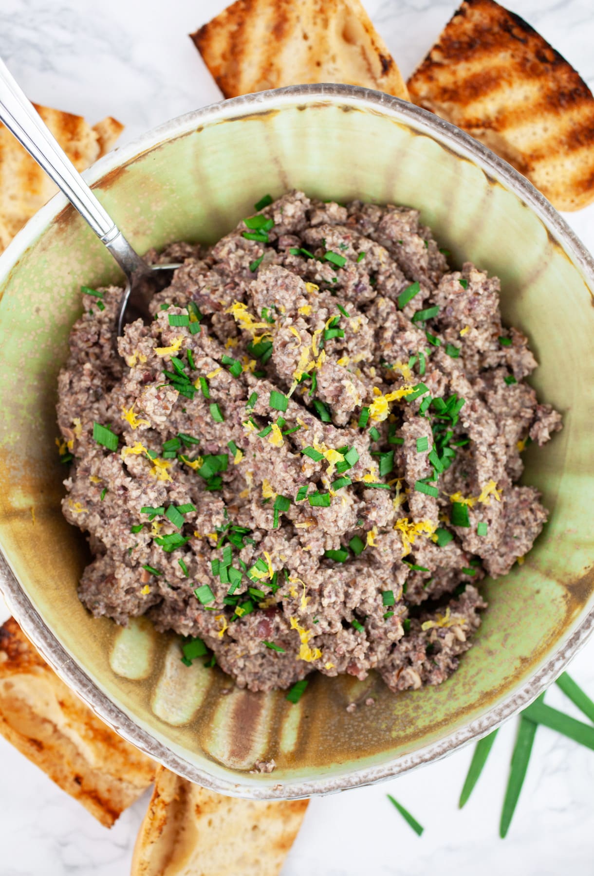 #AD MSG 4 21+ This Roasted Mushroom Tapenade Appetizer is the perfect simple and elegant recipe for your summer parties! Portobello and Crimini mushrooms are roasted on the grill and added to kalamata olives along with roasted garlic and walnuts. This healthy vegan and vegetarian dip is served on crostini. Serve with The Dreaming Tree wines for the perfect summer appetizer! #SeedOfAGreatSummer #mushrooms #tapenade #olives #roasted #appetizers Mushroom tapenade in ceramic bowl with spoon next to slices of toasted crostini.