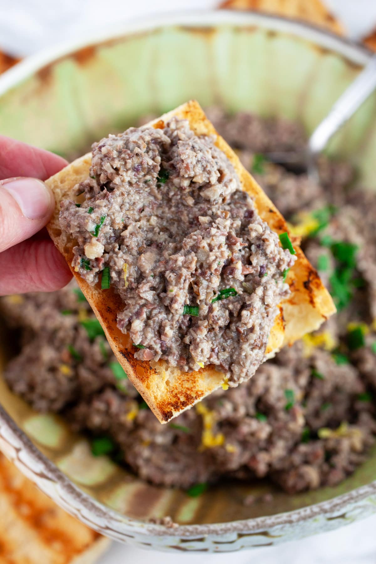 Mushroom olive tapenade on piece of toasted bread in front of ceramic bowl.