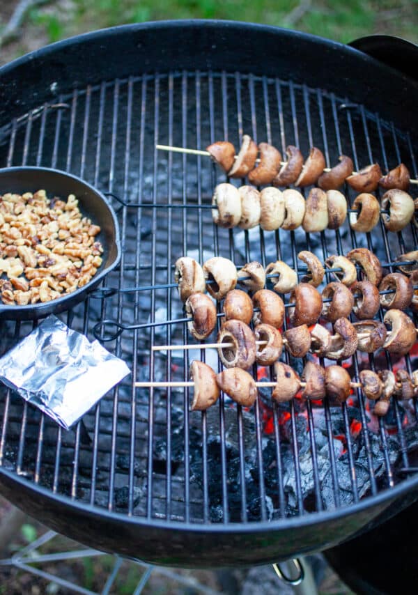 Mushroom skewers, tinfoil packet, and walnuts in small cast iron skillet cooking on grill.