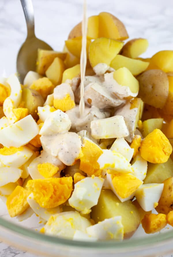 Creamy dressing poured onto cooked, diced potatoes and eggs in glass bowl with spoon.
