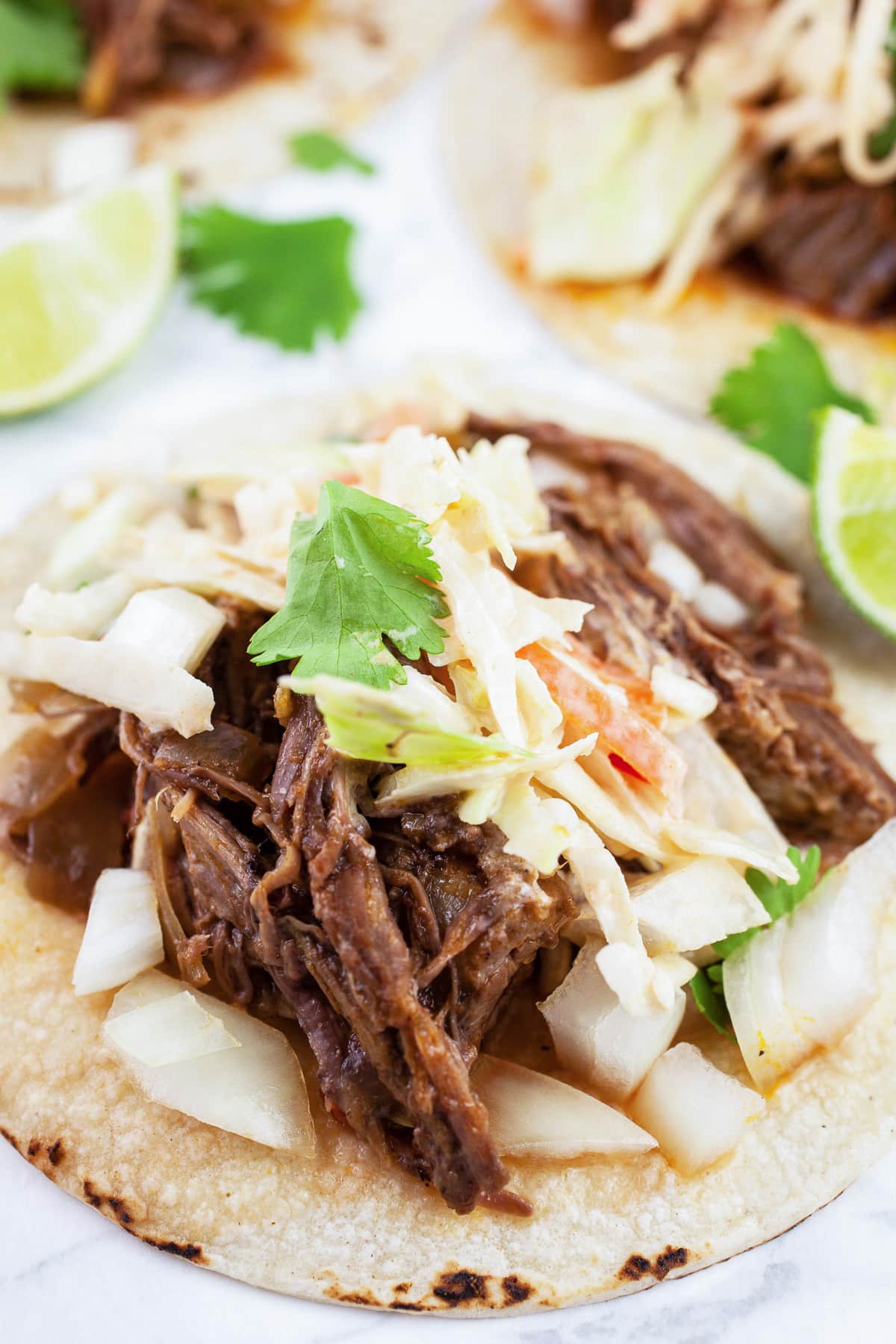 These Slow Cooker Barbacoa Tacos with Chipotle Slaw are full of flavorful shredded beef that's been slow cooked in the crockpot. They're topped with slaw that's flavored with chipotle in adobo. These tacos have tons of authentic Mexican spices. They're smoky and a little spicy. This easy dinner recipe is finished with fresh toppings like cilantro. One of my favorite beef recipes! #barbacoa #beef #slowcooker #crockpot #chipotle #withslaw #coleslaw Slow cooker barbacoa beef tacos with slaw on corn tortillas with onions, cilantro, and limes.