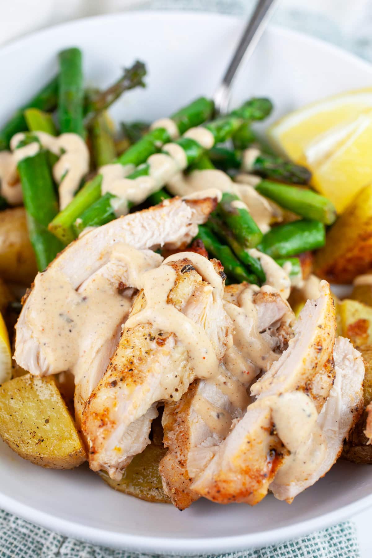 Roasted chicken thighs, potatoes, and asparagus topped with tahini sauce in white bowl with fork.