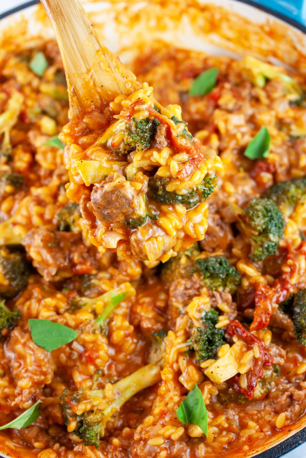 Wooden spoonful of sausage broccoli pasta lifted from skillet.