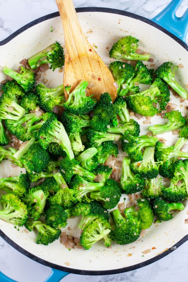 Broccoli florets sauteed in skillet with wooden spoon.
