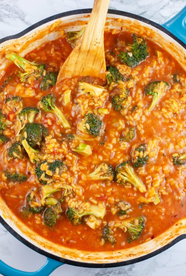 Broccoli and orzo pasta cooked in tomato sauce in skillet with wooden spoon.