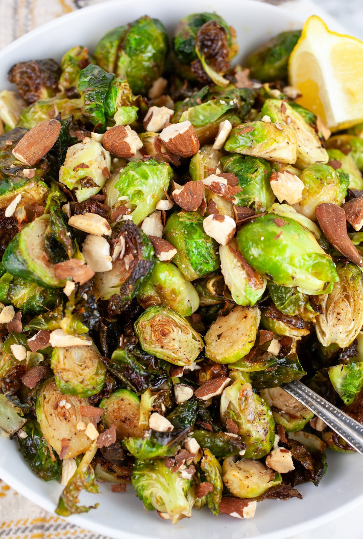 Roasted maple mustard Brussels sprouts in white bowl with almonds and wedge of lemon with spoon.