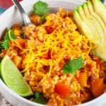 Cheesy Spanish rice in white bowl with fork garnished with cilantro, avocado slices, and lime wedge.