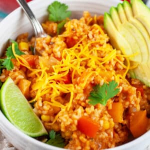 Cheesy Spanish rice in white bowl with fork garnished with cilantro, avocado slices, and lime wedge.