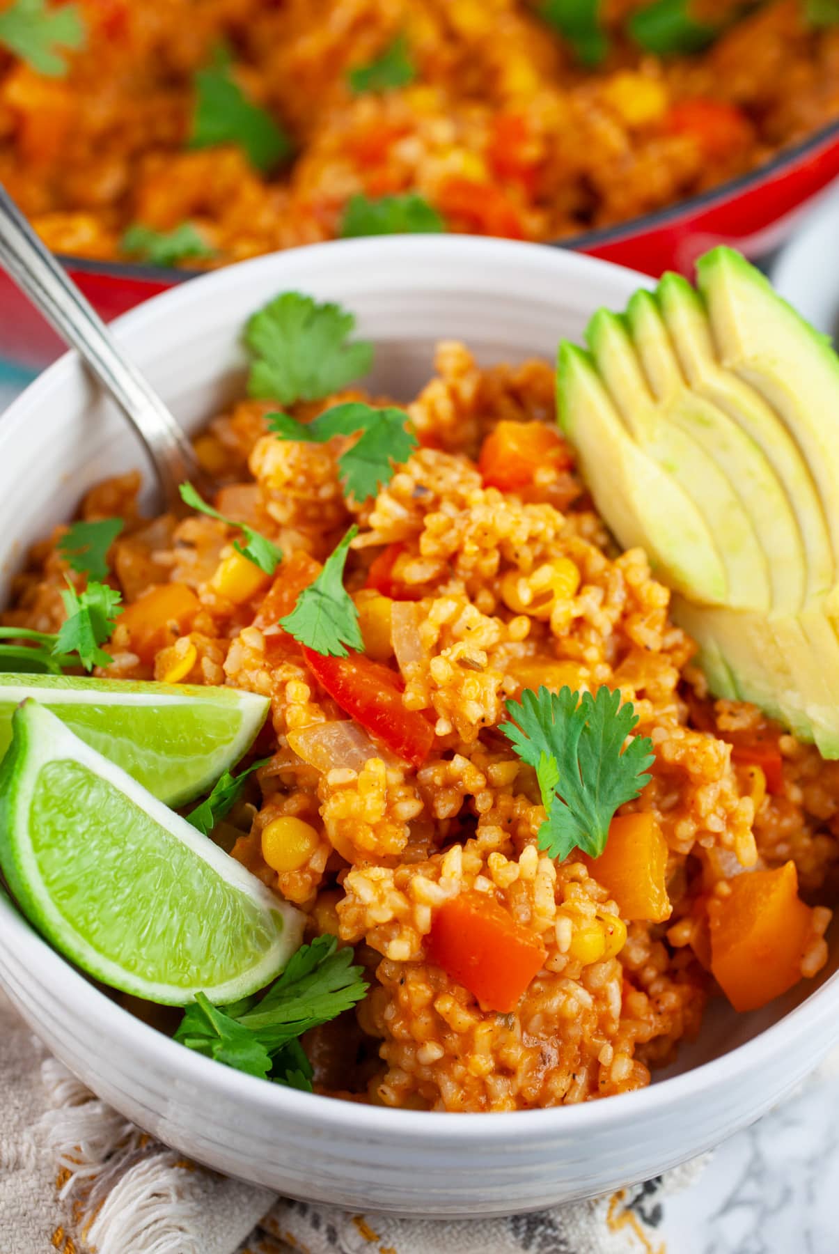 This Cheesy One Pot Spanish Rice is a quick and easy side dish to make for dinner! This homemade Spanish rice is authentic, healthy, and vegetarian. It's made with tomato sauce and cooks on the stove top. The perfect side dish for taco night! #ad #CheeseLove @crystalfarmscheese #onepot #spanishrice #vegetarian Spanish rice in white bowl with fork garnished with cilantro, limes wedges, and sliced avocado.