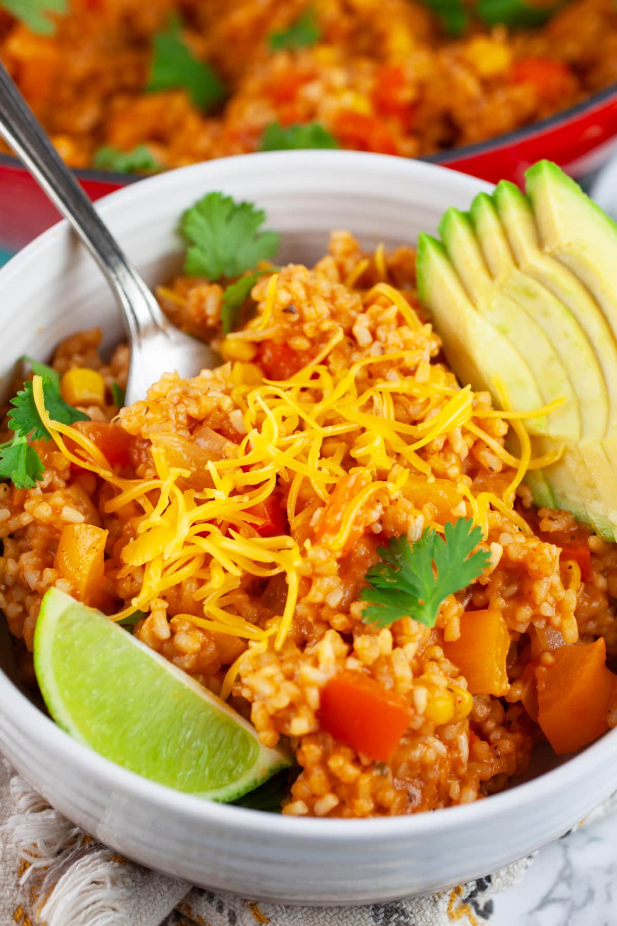 This Cheesy One Pot Spanish Rice is a quick and easy side dish to make for dinner! This homemade Spanish rice is authentic, healthy, and vegetarian. It's made with tomato sauce and cooks on the stove top. The perfect side dish for taco night! #ad #CheeseLove @crystalfarmscheese #onepot #spanishrice #vegetarian Cheesy Spanish rice in white bowl with fork garnished with fresh cilantro, sliced avocado, and lime wedge.