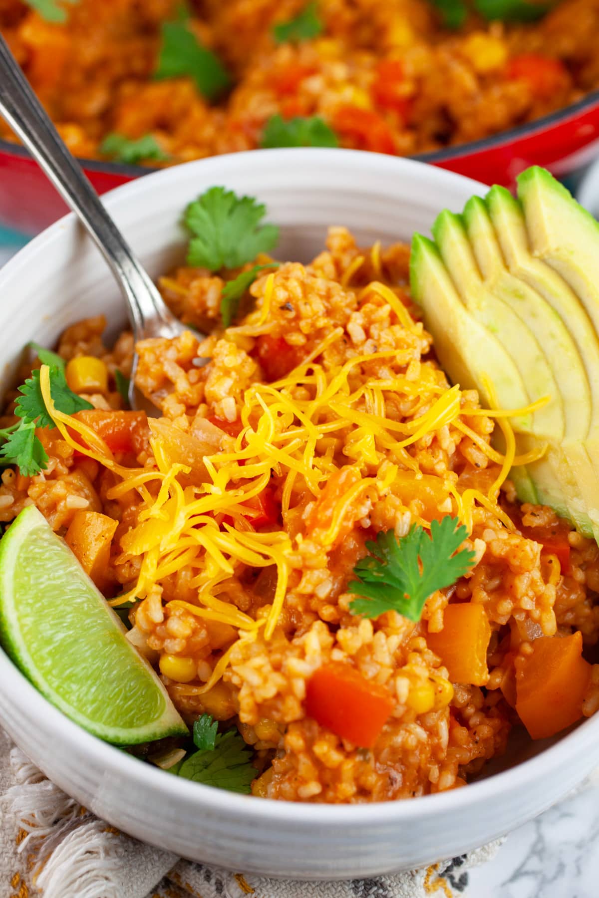 This Cheesy One Pot Spanish Rice is a quick and easy side dish to make for dinner! This homemade Spanish rice is authentic, healthy, and vegetarian. It's made with tomato sauce and cooks on the stove top. The perfect side dish for taco night! #ad #CheeseLove @crystalfarmscheese #onepot #spanishrice #vegetarian Cheesy Spanish rice in white bowl with fork garnished with cilantro, lime wedge, and avocado slices.