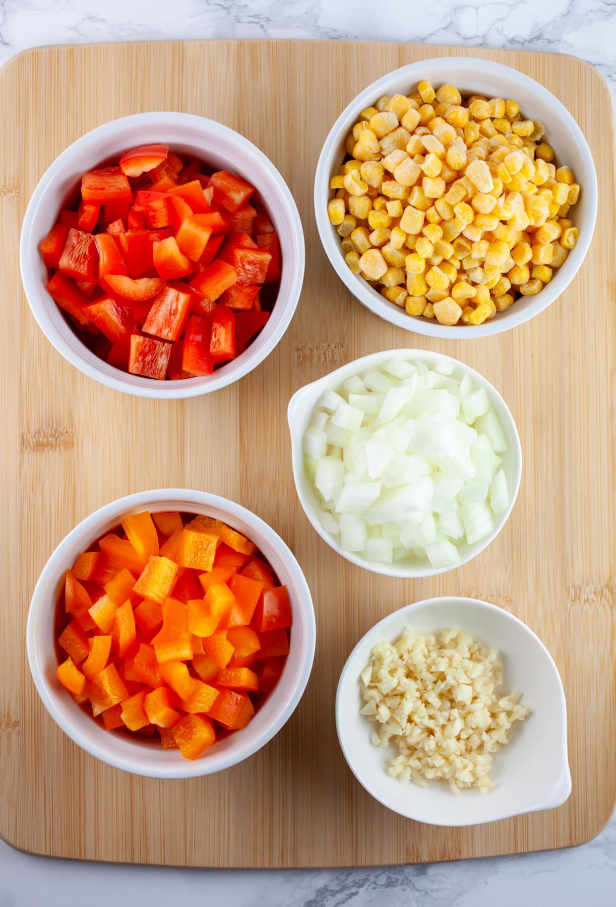 This Cheesy One Pot Spanish Rice is a quick and easy side dish to make for dinner! This homemade Spanish rice is authentic, healthy, and vegetarian. It's made with tomato sauce and cooks on the stove top. The perfect side dish for taco night! #ad #CheeseLove @crystalfarmscheese #onepot #spanishrice #vegetarian Minced garlic, onions, red and orange bell peppers, and sweet corn kernels in small white bowls on wooden cutting board.