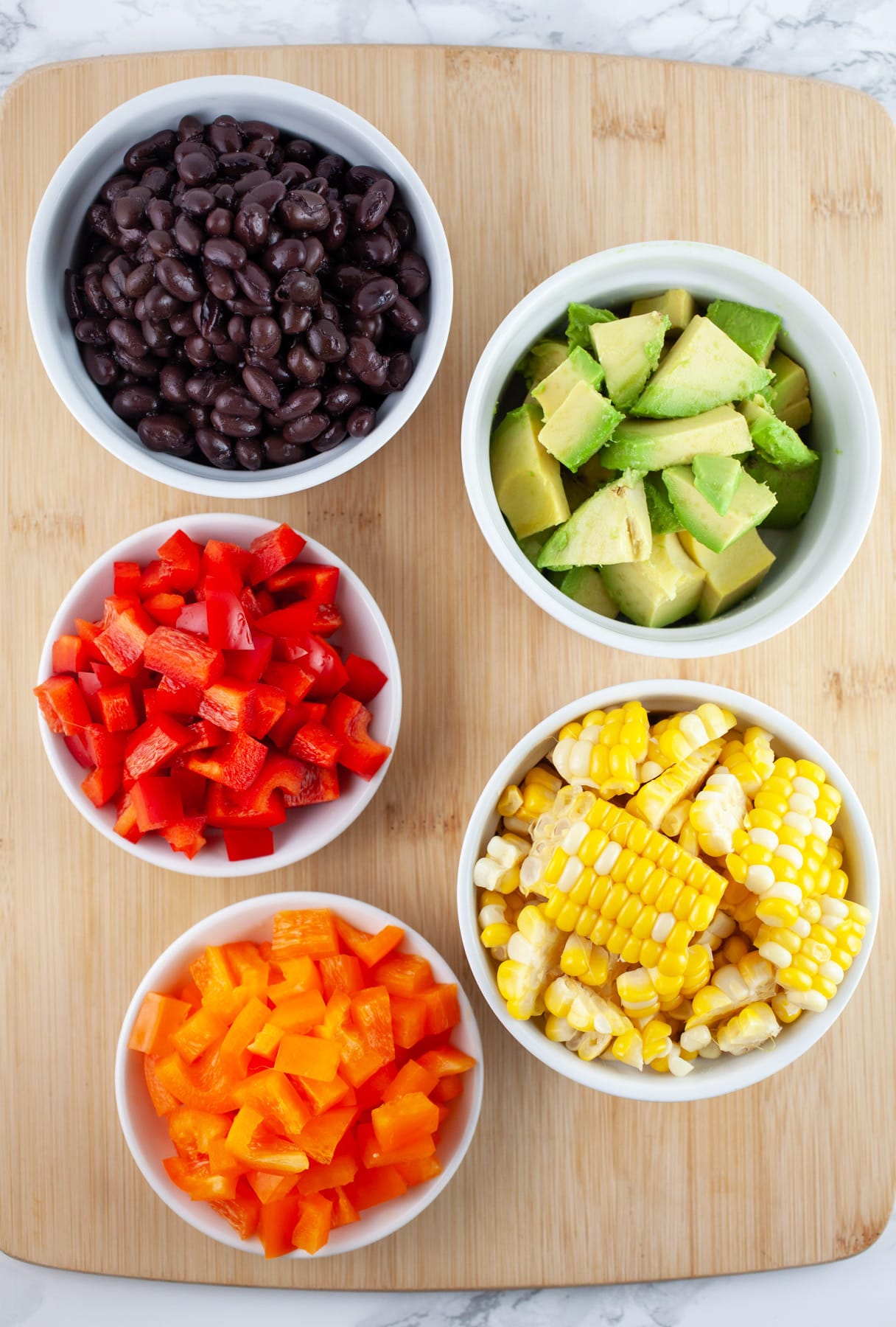 Sweet corn, diced orange and red bell peppers, black beans, and avocadoes in white bowls on wooden cutting board.