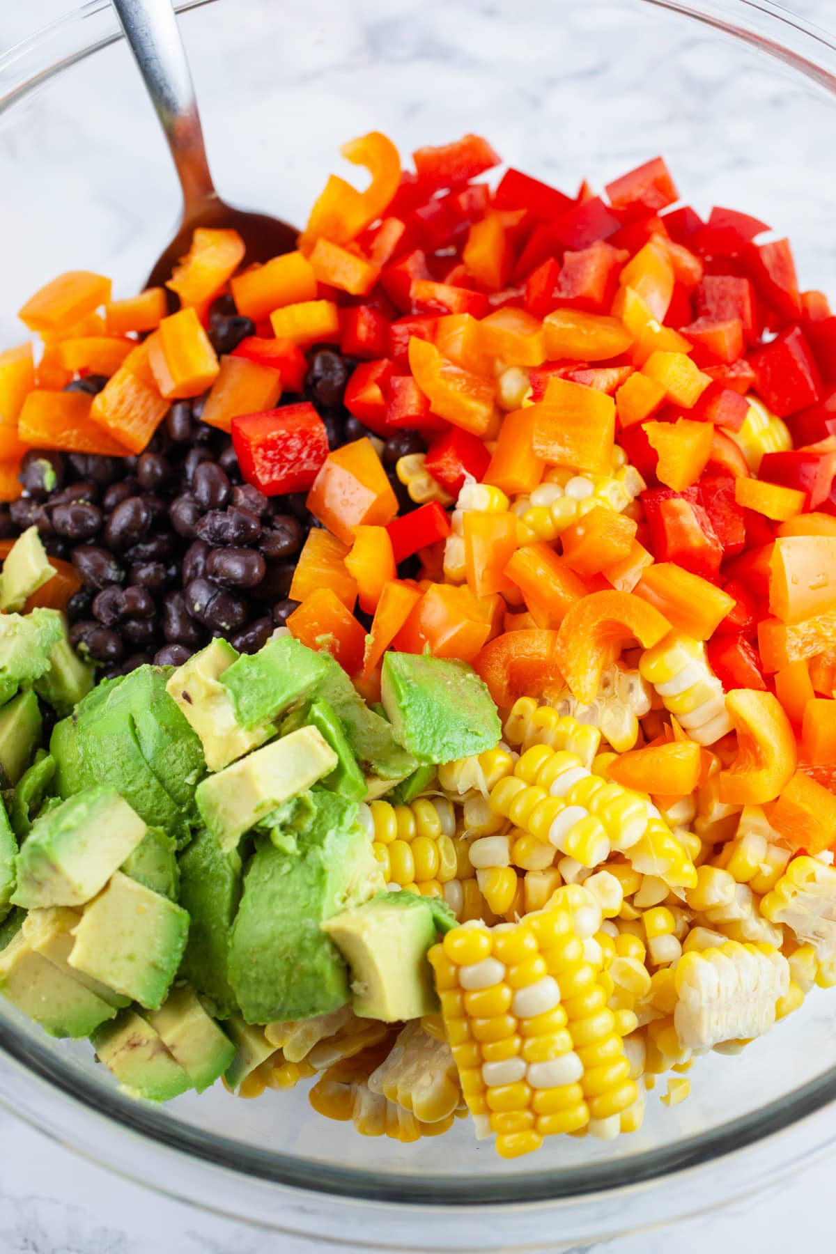 Sweet corn, diced red and orange bell peppers, black beans, and diced avocado in large glass bowl with spoon.