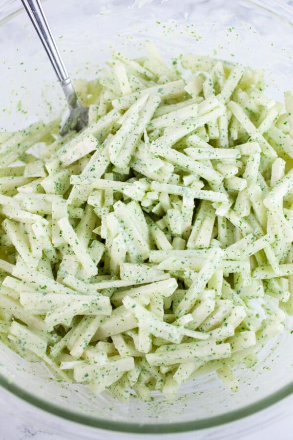 Sliced kohlrabi tossed in cilantro lime dressing in glass bowl with spoon.