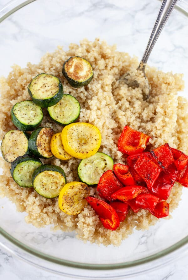 Cooked quinoa, roasted zucchini and summer squash, and red bell peppers in glass mixing bowl with spoon.