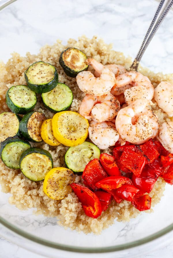 Cooked quinoa, roasted zucchini and summer squash, red bell peppers, and shrimp in glass mixing bowl with spoon.