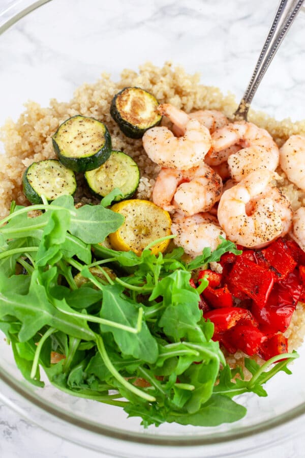 Cooked quinoa, roasted zucchini and summer squash, red bell peppers, and fresh arugula in glass mixing bowl with spoon.