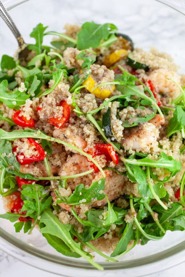 Quinoa zucchini shrimp arugula salad in glass mixing bowl with spoon.