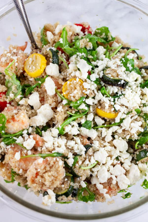 Quinoa shrimp zucchini salad with feta cheese in glass mixing bowl with spoon.