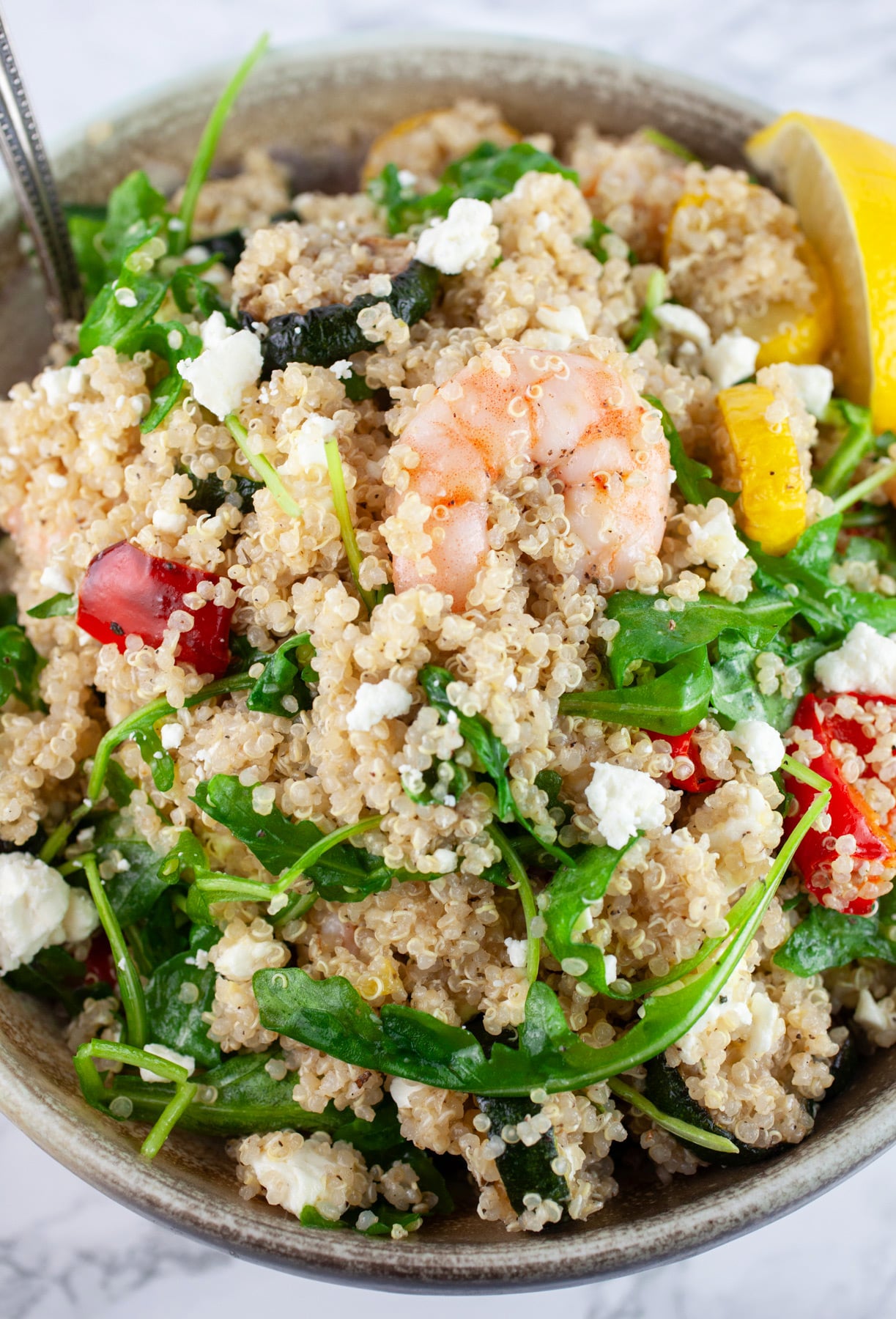 Quinoa zucchini shrimp salad with arugula garnished with lemon wedge in ceramic bowl with spoon.
