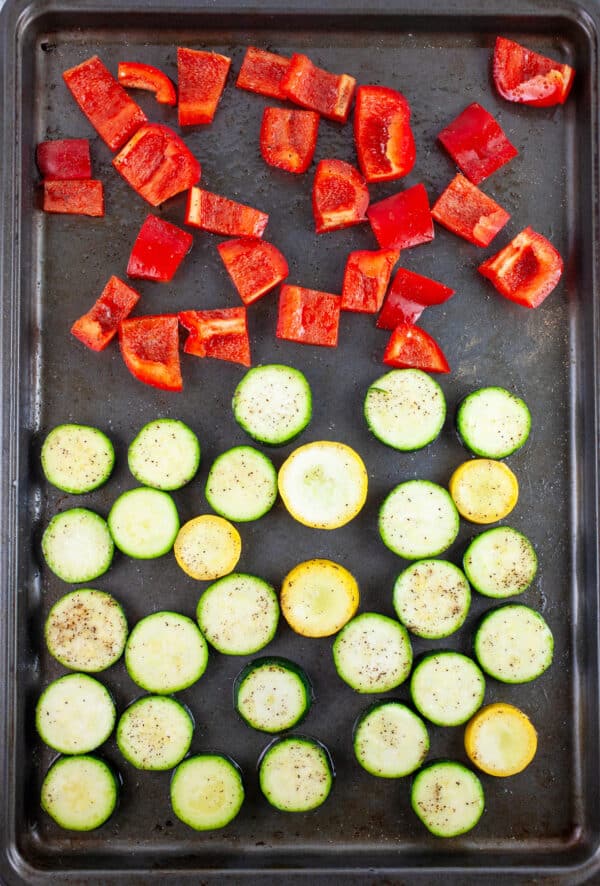 Chopped zucchini, summer squash, and red bell peppers tossed in olive oil, salt and pepper on baking sheet.