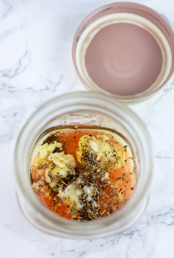 Grated garlic, olive oil, red wine vinegar, Dijon mustard, Italian seasoning, and salt and pepper in small mason jar sitting next to lid.