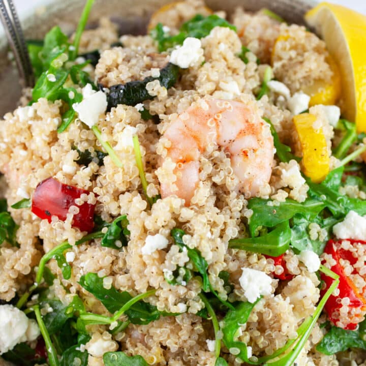 Quinoa salad with shrimp, vegetables, arugula, and feta cheese in bowl garnished with lemon wedge.