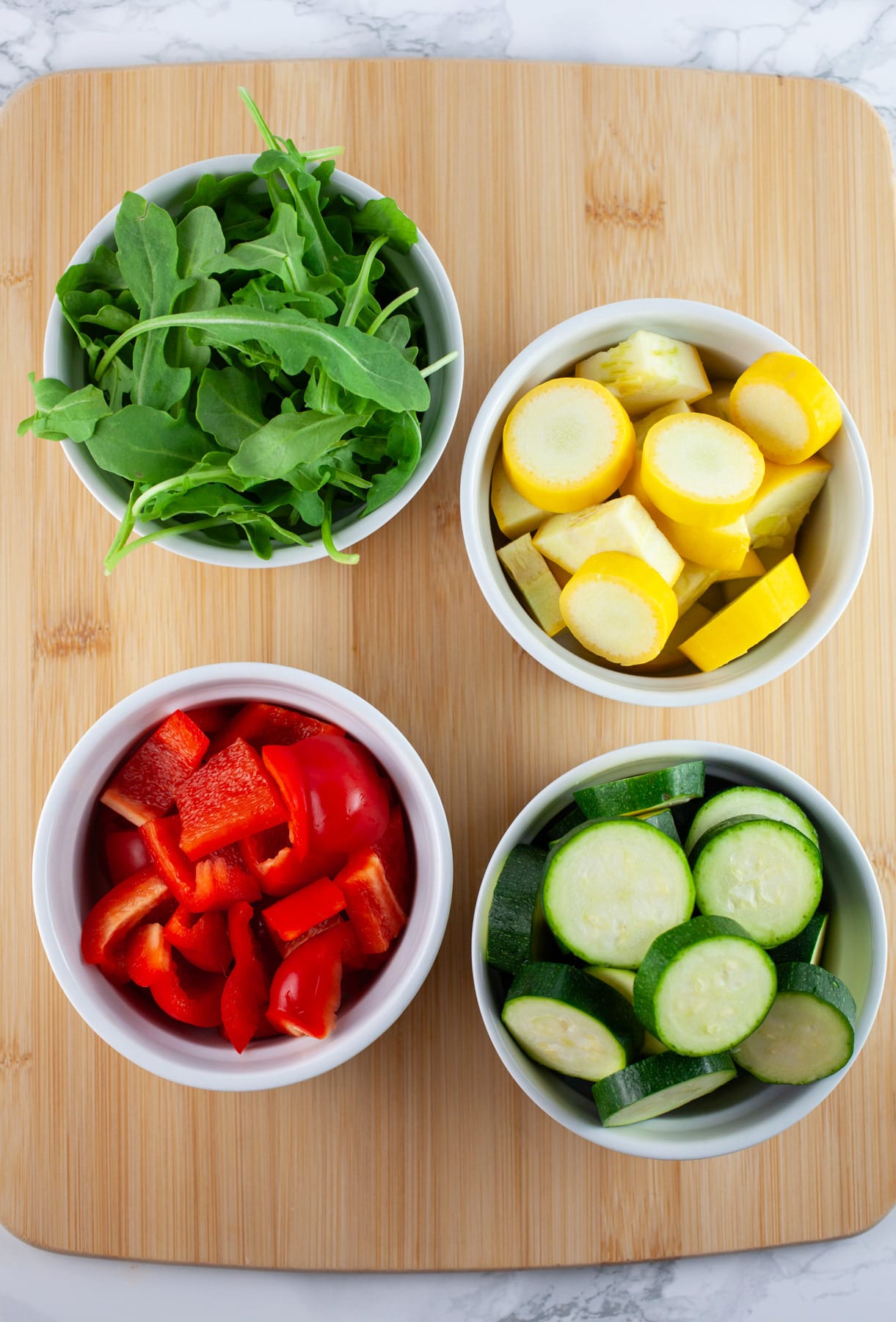 Chopped zucchini, summer squash, and red bell peppers and arugula in small white bowls on wooden cutting board.