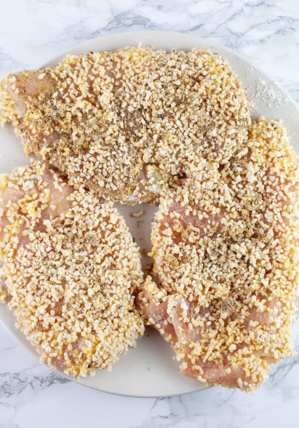 Raw chicken breasts coated in spiced Panko breadcrumbs on white plate.