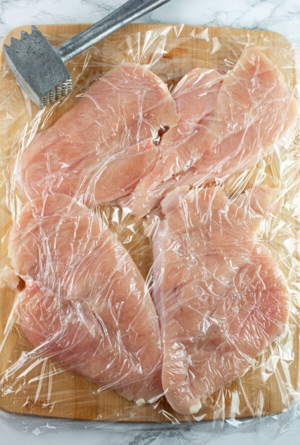 Raw chicken breasts cut in half covered with plastic wrap on wooden cutting board with meat tenderizer.