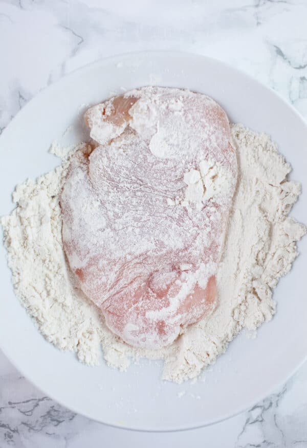 Raw chicken breast coated in flour in white bowl.
