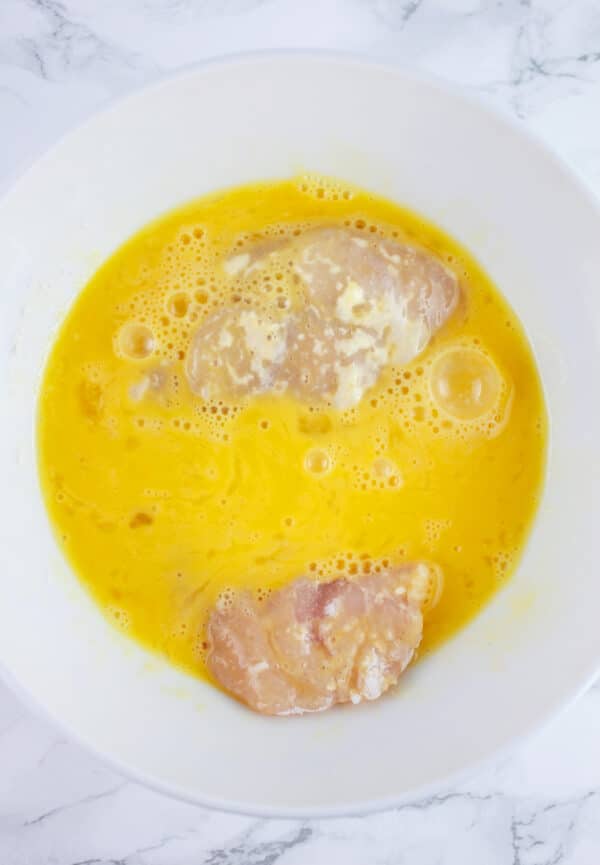 Raw chicken breasts coated in egg mixture in white bowl.