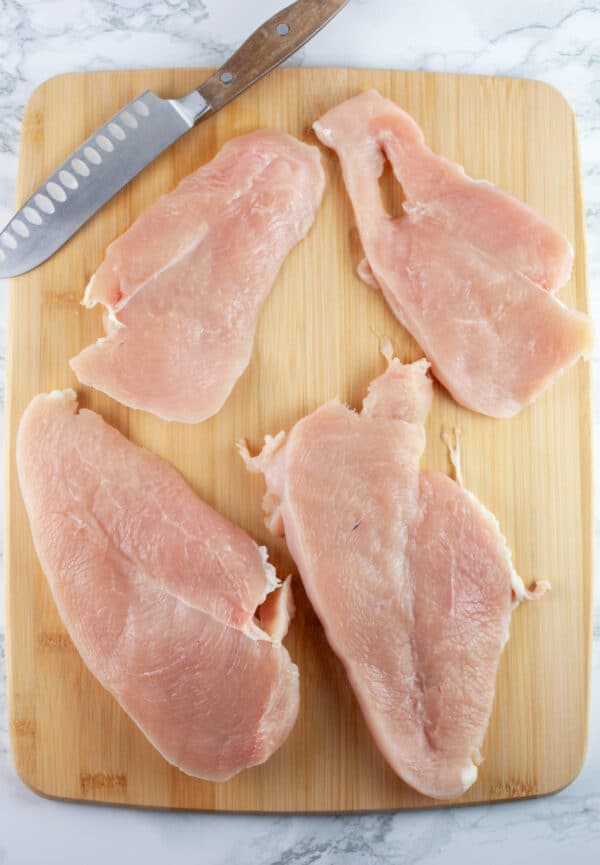 Raw chicken breasts cut in half on wooden cutting board with knife.