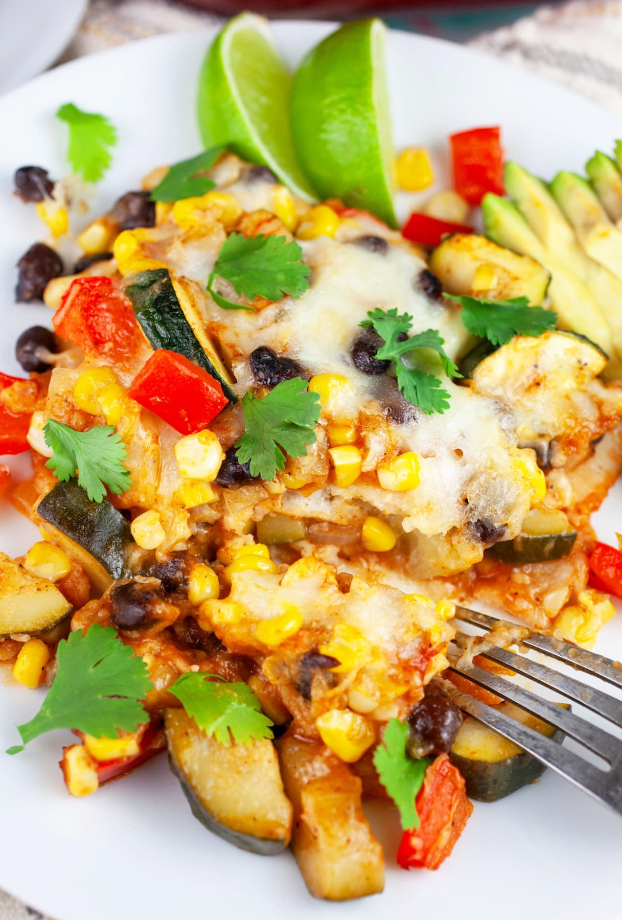 Vegetarian enchilada casserole on white plate with fork garnished with avocado slices, lime wedges, and fresh cilantro.