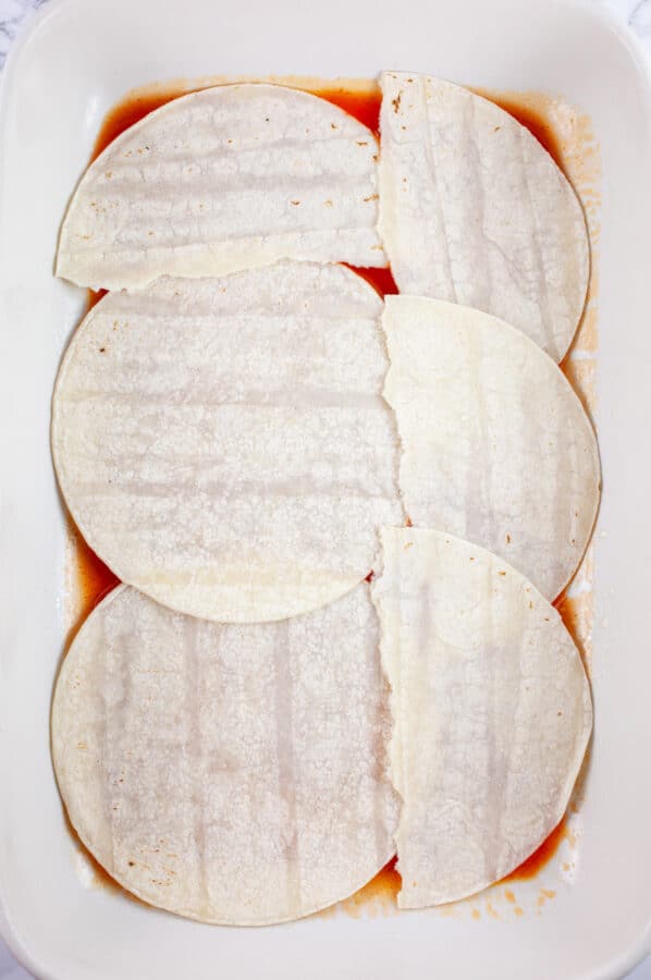 Corn tortillas and red enchilada sauce in ceramic casserole dish.