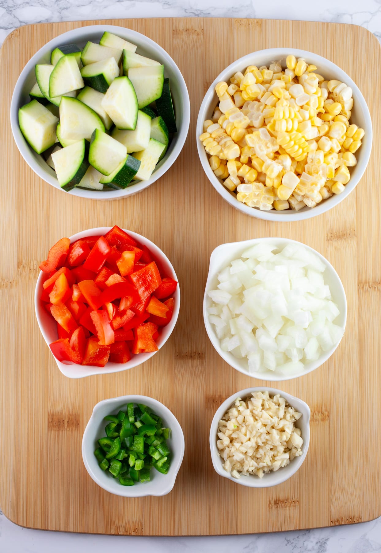 Minced garlic, onions, jalapenos, diced red bell peppers and zucchini, and sweet corn in small white bowls on wooden cutting board.