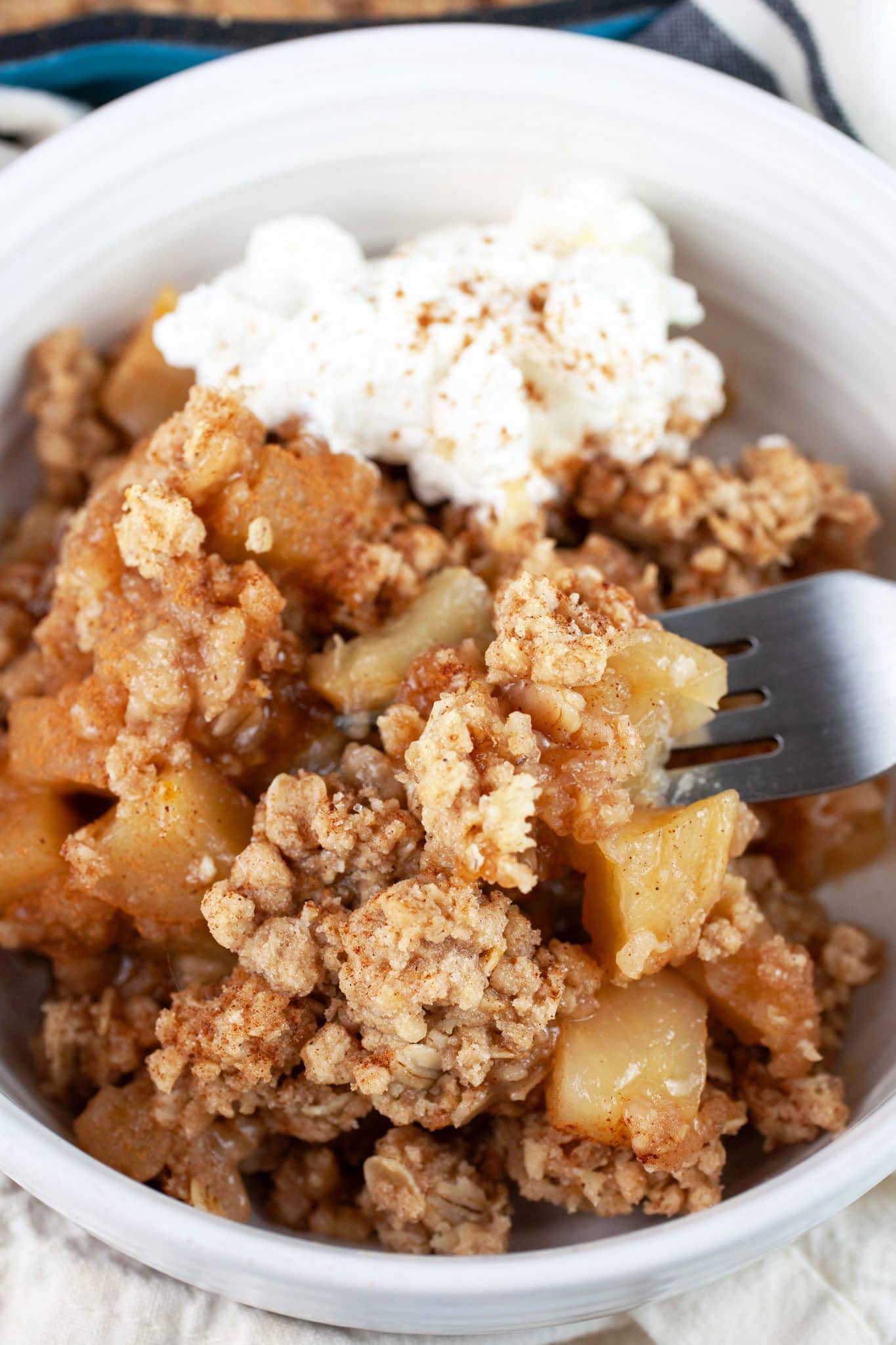 Apple crisp in white bowl with fork with homemade whipped cream.